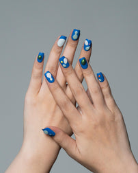 A close-up of two hands crossed together, showcasing a manicure with blue almond-shaped nails decorated with white abstract spots and one green accent nail, set against a solid gray background.
