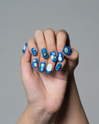 A close-up of a hand with manicured fingernails painted in a blue abstract pattern with white, light blue, and small green accents, set against a solid gray background.