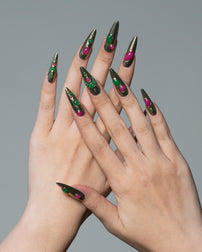A pair of hands with long, sharp stiletto-shaped nails painted in a metallic olive green base with glittery green and magenta accents, posed against a solid gray background.