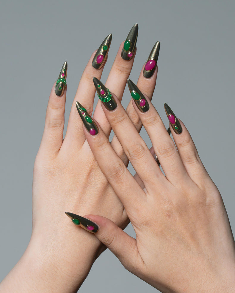 A pair of hands with long, sharp stiletto-shaped nails painted in a metallic olive green base with glittery green and magenta accents, posed against a solid gray background.