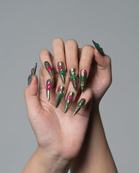 A pair of hands with long stiletto-shaped nails decorated in a metallic olive green and silver pattern with bright pink accents, set against a solid gray background.