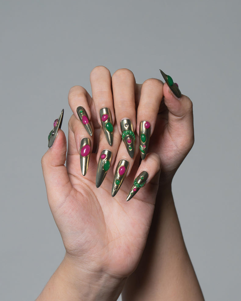A pair of hands with long stiletto-shaped nails decorated in a metallic olive green and silver pattern with bright pink accents, set against a solid gray background.