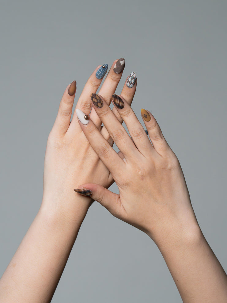 Two hands crossed together showcasing a set of long almond-shaped press-on nails with various designs including brown animal prints, blue plaid, and white patterns, set against a solid gray background.
