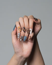 A close-up of two hands clasped together, showcasing a set of long almond-shaped nails with various autumn-themed patterns including plaid, argyle, checkered, and solid brown, beige, and blue designs, set against a solid gray background.