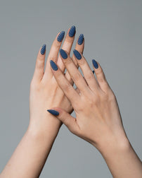 A pair of hands with long, almond-shaped nails painted in a dark slate blue color with small white polka dots, shown against a solid gray background.