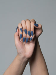A close-up of a person's hands with long, almond-shaped nails painted in a dusty slate blue color, decorated with small white polka dots, set against a solid gray background.