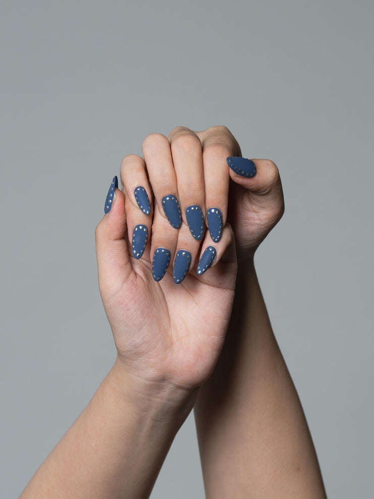 A close-up of a person's hands with long, almond-shaped nails painted in a dusty slate blue color, decorated with small white polka dots, set against a solid gray background.