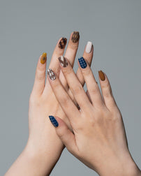 A pair of hands with styled almond-shaped nails featuring a mix of brown animal prints, blue plaid, white, and mustard yellow, set against a solid gray background.