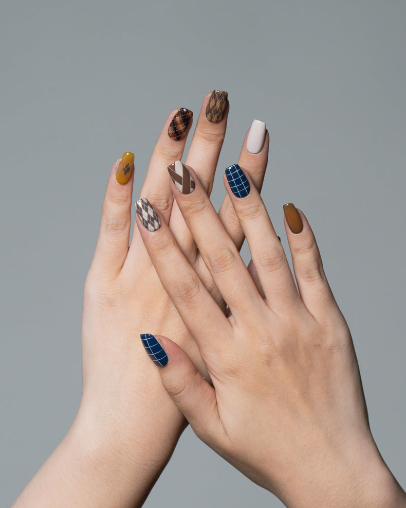 A pair of hands with styled almond-shaped nails featuring a mix of brown animal prints, blue plaid, white, and mustard yellow, set against a solid gray background.