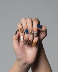 A close-up of a person's hands showcasing a fall-themed nail art set with brown, beige, blue, and white patterns against a solid gray background.