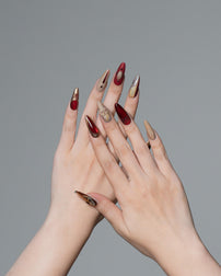 A pair of hands with long, sharp stiletto-shaped nails painted in alternating deep red and metallic gold, posed against a solid gray background.