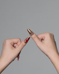 Two hands against a solid gray background, showcasing long, sculpted stiletto-shaped nails. One nail features a metallic gold base with a silver glittery pattern, and the other features a glossy deep red finish.