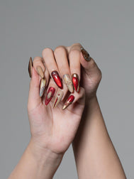 A pair of hands with long, sculpted stiletto-style nails decorated in a metallic red, gold, and beige pattern, posed against a solid gray studio background.