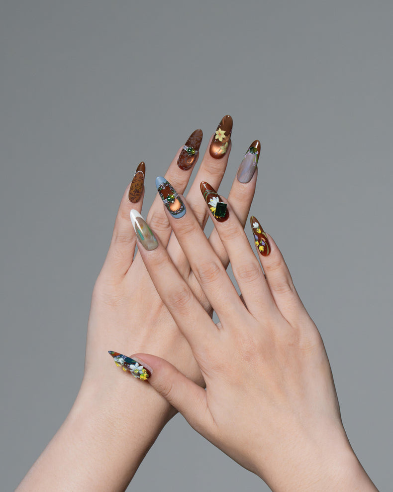 A pair of hands with long, almond-shaped stiletto nails decorated with intricate metallic and multicolor nail art, shown against a solid gray background.