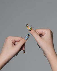 A close-up of two hands against a solid gray background, showcasing long, sculpted almond-shaped press-on nails with colorful, detailed floral and geometric patterns.