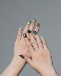 A pair of hands with manicured nails painted in various green shades, including solid olive green, metallic forest green, dark green, and white patterns, shown against a solid gray background.