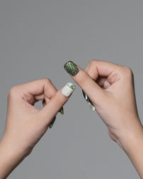 Two hands holding a single long artificial nail against a plain gray background. The nail has a dark green glittery top with a white glittery accent band and small green leaf-like details near the cuticle.