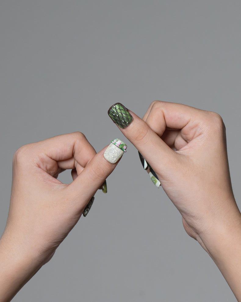 Two hands holding a single long artificial nail against a plain gray background. The nail has a dark green glittery top with a white glittery accent band and small green leaf-like details near the cuticle.