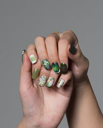 A close-up of two hands with manicured fingernails, showcasing a variety of nail art designs including green glitter, floral patterns, and solid olive green, set against a solid gray background.