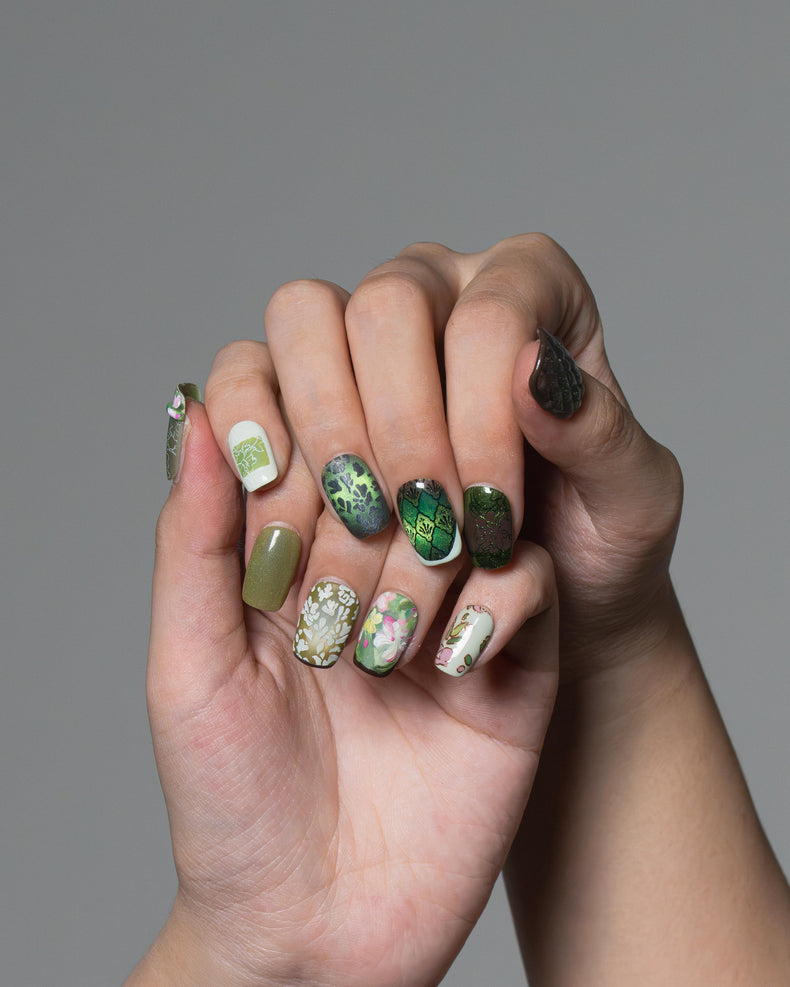 A close-up of two hands with manicured fingernails, showcasing a variety of nail art designs including green glitter, floral patterns, and solid olive green, set against a solid gray background.