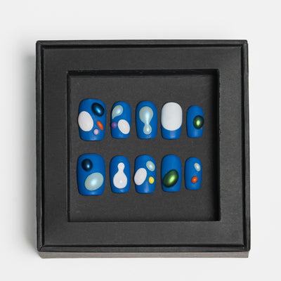A set of 10 blue press-on nails with abstract white, green, and orange bubble patterns, displayed inside a square black frame on a light gray background.