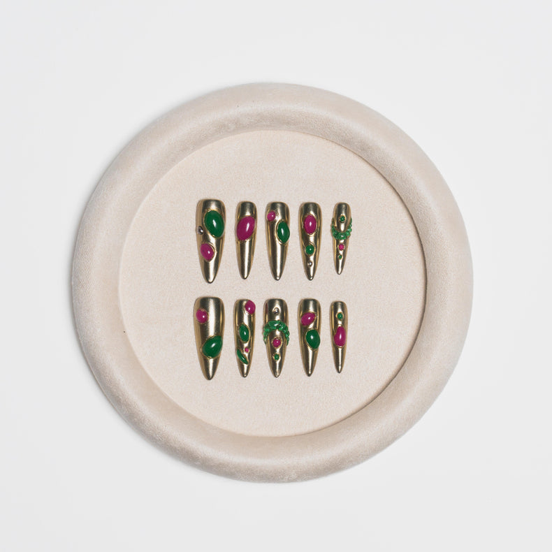 A set of metallic gold press-on nails with green and pink floral details, displayed on a round beige velvet ring stand against a light gray background.