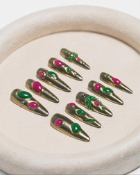 Nine long metallic gold coffin-shaped press-on nails with green and pink gemstone embellishments, arranged on a beige textured circular tray.