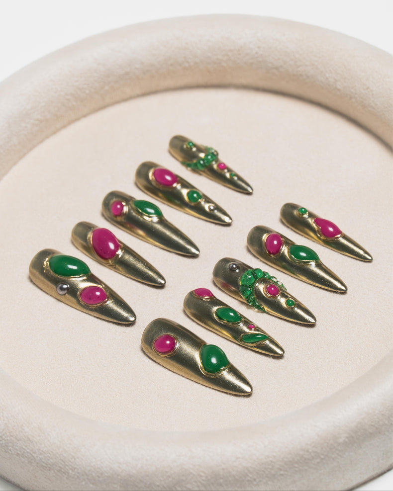 Nine long metallic gold coffin-shaped press-on nails with green and pink gemstone embellishments, arranged on a beige textured circular tray.