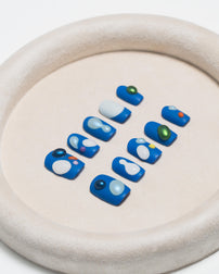 A set of blue press-on nails with white, light blue, and colorful bead accents, arranged on a beige textured circular tray.