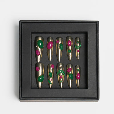 A set of 10 coffin-shaped press-on nails with a metallic gold base, decorated with green and pink faux gemstones, presented in a black square box on a light gray background.