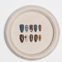 A circular beige display tray holding ten artificial press-on nails in various patterns and colors, including blue plaid, brown argyle, solid brown, and beige with a pearl accent, set against a plain white background.