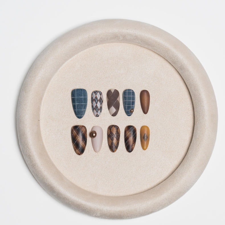 A circular beige display tray holding ten artificial press-on nails in various patterns and colors, including blue plaid, brown argyle, solid brown, and beige with a pearl accent, set against a plain white background.