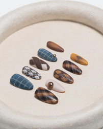A set of ten press-on nails arranged in a circle on a textured beige tray. The nails feature various patterns including plaid, argyle, stripes, and solid colors in shades of brown, blue, beige, and yellow.