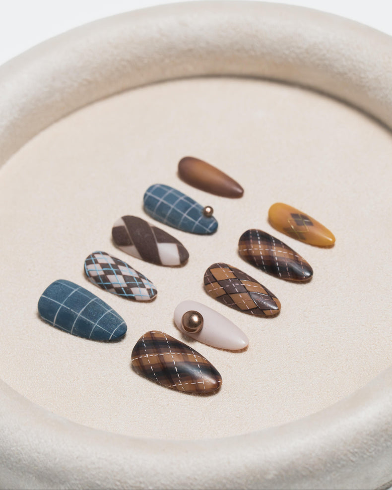 A set of ten press-on nails arranged in a circle on a textured beige tray. The nails feature various patterns including plaid, argyle, stripes, and solid colors in shades of brown, blue, beige, and yellow.