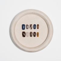A circular beige tray displaying ten press-on nails in various patterns and colors, including plaid, marble, solid brown, and neutral shades, set against a clean white background.