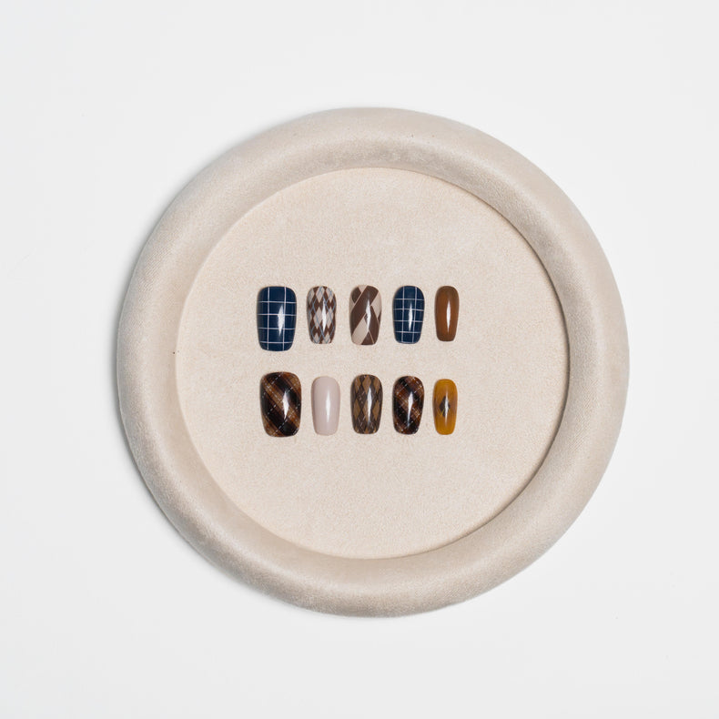 A circular beige tray displaying ten press-on nails in various patterns and colors, including plaid, marble, solid brown, and neutral shades, set against a clean white background.