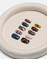 A set of ten press-on nails arranged on a round beige tray. The nails feature various patterns including plaid, argyle, stripes, and solid colors in shades of brown, navy, beige, and mustard yellow.