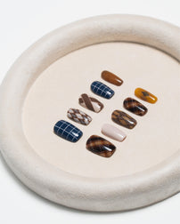 A set of ten press-on nails with various patterns including plaid, checkered, solid brown, and tiger stripe, arranged on a beige textured circular tray against a white background.
