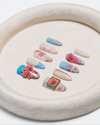 A set of decorative press-on nails arranged on a round beige tray. The nails feature pastel blue, pink, and nude tones with floral prints, 3D pink flowers, glitter, and one small gold-framed floral accent.