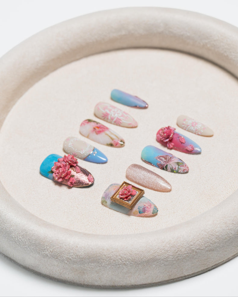 A set of decorative press-on nails arranged on a round beige tray. The nails feature pastel blue, pink, and nude tones with floral prints, 3D pink flowers, glitter, and one small gold-framed floral accent.