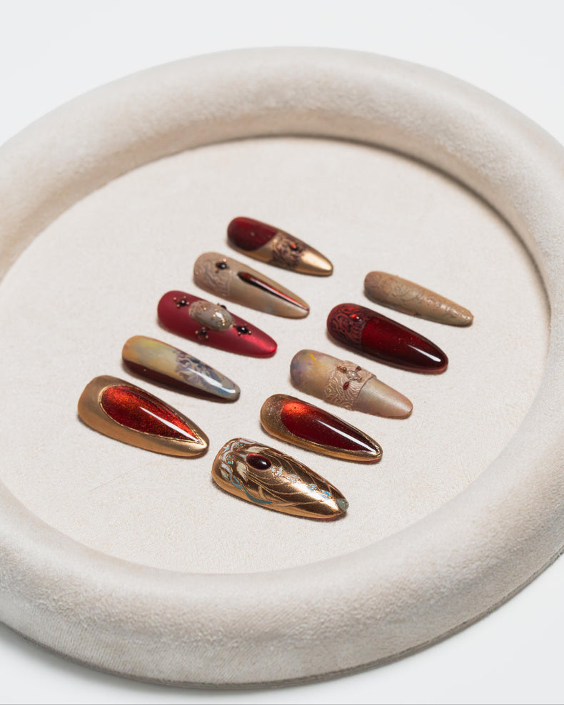Nine long coffin-shaped press-on nails with metallic gold, bronze, and deep red designs, displayed on a textured beige ceramic dish against a white background.
