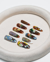 Nine decorative press-on nails arranged on a round beige tray against a white background. The nails feature glossy brown, blue-gray, and dark marbled designs with floral and leaf embellishments in yellow, white, green, and pink.