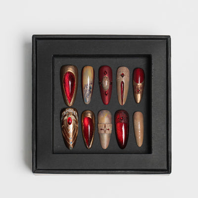 A set of 10 coffin-shaped press-on nails displayed in a black square tray against a light gray background. The nails feature a mix of metallic gold, deep red, and textured beige designs.