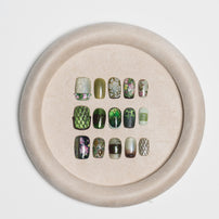 A set of 15 false press-on nails displayed on a beige circular stand against a light gray background. The nails feature various designs including green snakes, floral patterns, and textured prints.