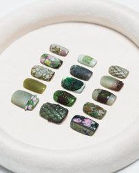 A set of 15 press-on nails with various designs including floral prints, geometric patterns, and solid colors, arranged on a round beige velvet tray.